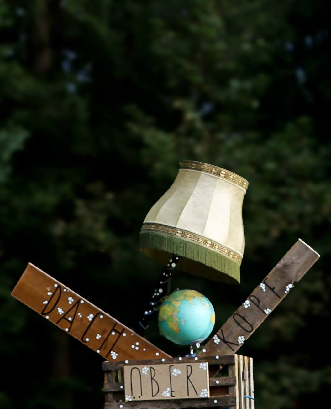 Das DachüberKopf Installationskunst-Element: gestapelte Holzkisten mit Globus und Lampenschirm, umgeben von Wald. Kunstinstallation auf dem Festivalgelände Moosach 2025.